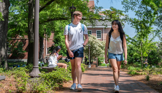 Two undergraduate students walk down a brick path near the base of Tech Tower. In the background, greenery and other students are visible, as well as historic brick buildings.